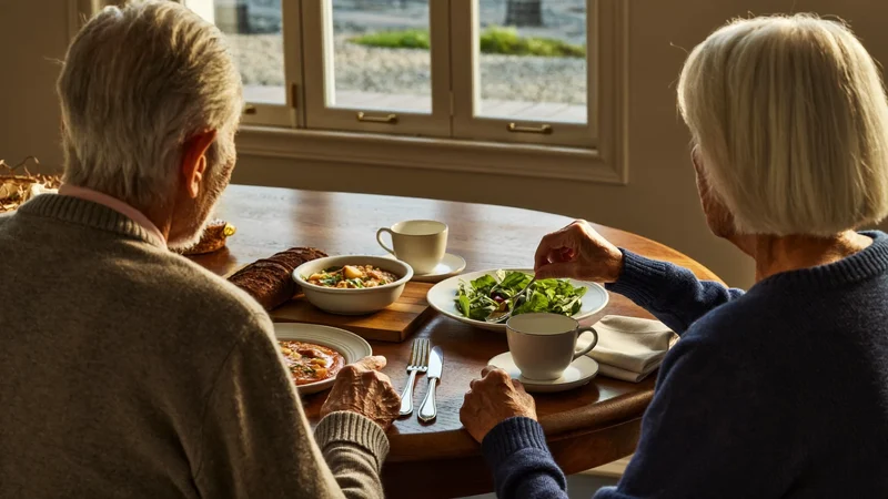 Zwei ältere Menschen, von hinten, beim gemeinsamen Essen am Holztisch am Fenster